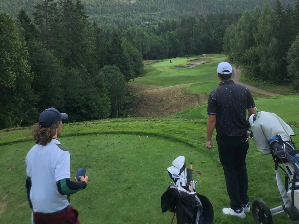 To golfspillere med golf bags på teestedet med skovklædte bjerge bag sig
