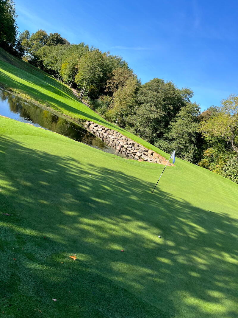 Skrånende fairway langs en vandhindring med stensat kant og træer